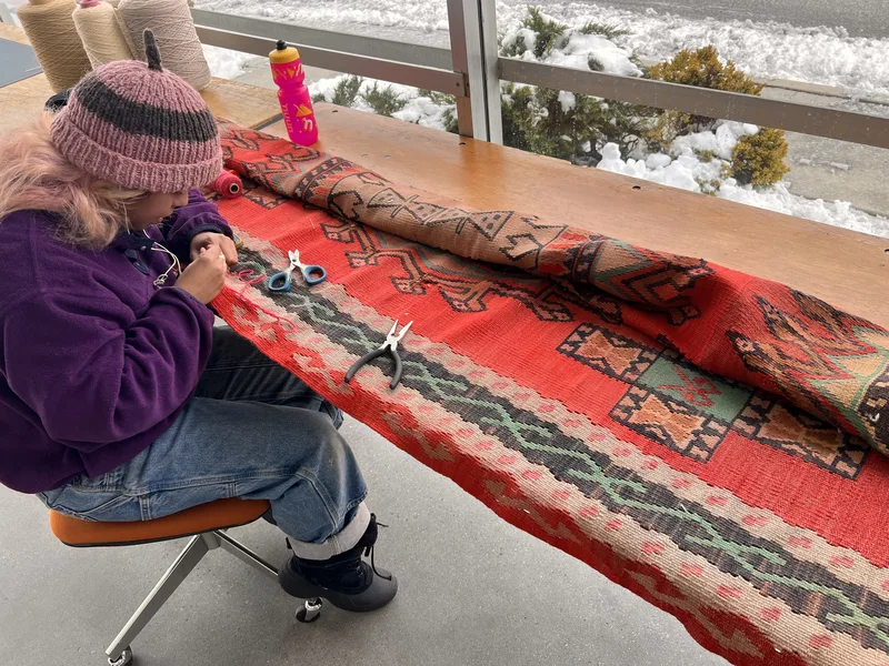 A repair specialist hand sewing slitweave damage on a red Turkish kilim at the Fiber Specialties Boise shop